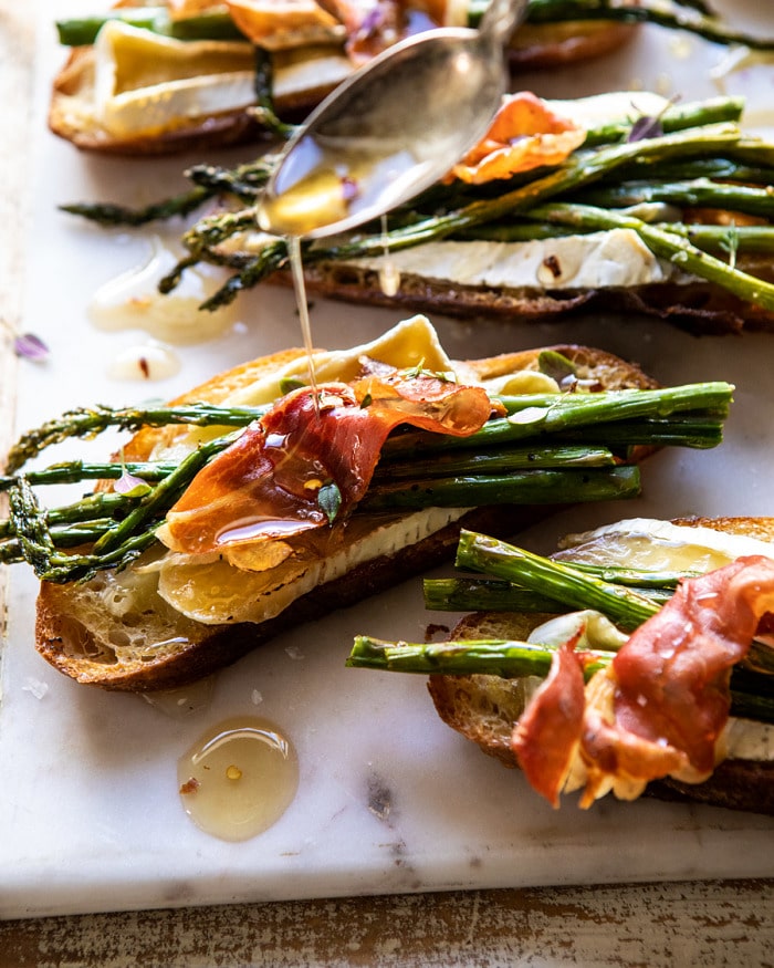 side angled photo of Crispy Prosciutto, Asparagus, and Brie Toast with honey being drizzled over the toast