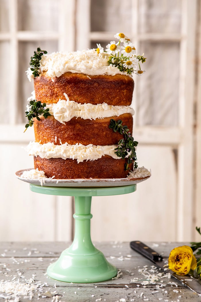side angled photo of Lemon Coconut Naked Cake with Whipped Vanilla Buttercream on cake stand and flowers on table