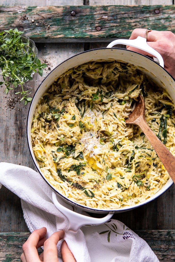 overhead photo of Souper Creamy Lemon Butter Cheesy Zucchini Orzo with hand on pot 