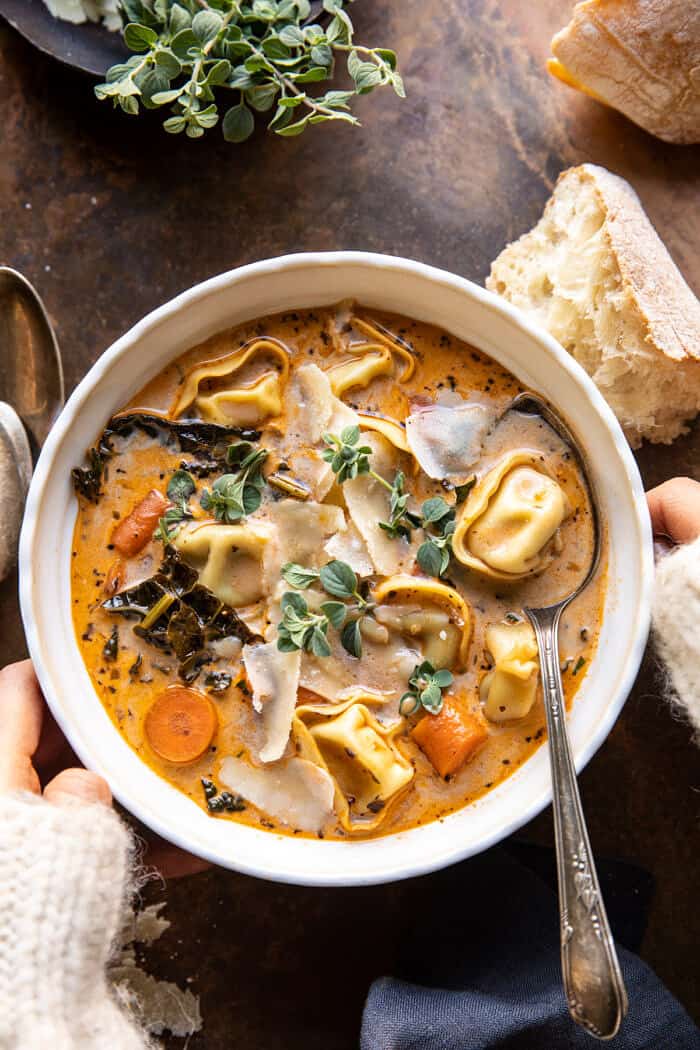 overhead photo of Healthier Slow Cooker Creamy Tortellini Vegetable Soup with 2 hands on bowl